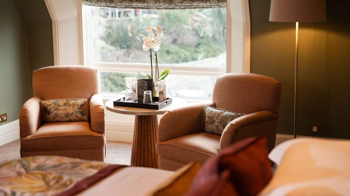 Two beige armchairs with patterned cushions sit around a small round table holding a tray with glassware and a potted orchid, next to a large window in a softly lit room.