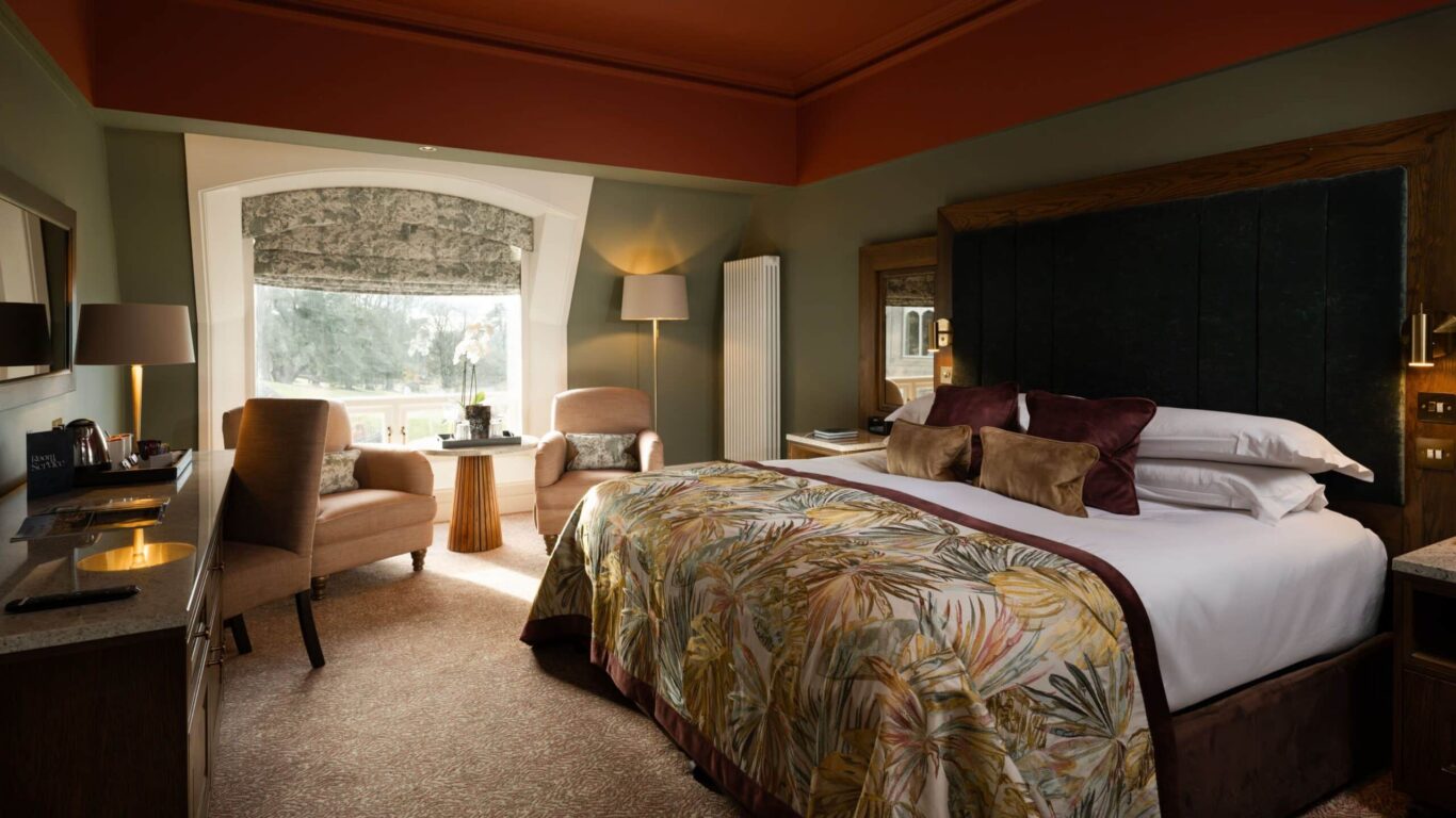 A neatly arranged hotel room with a king-size bed, two armchairs by a large window, a desk, and warm, muted decor.