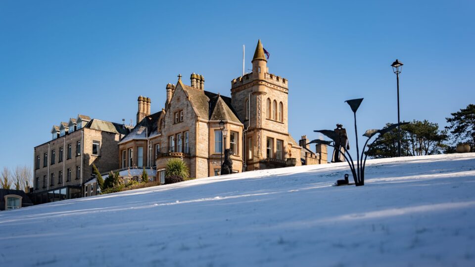 A large stone building with a tower stands on a snow-covered hill under a clear blue sky. Modern street lamps and some trees are visible in the scene.