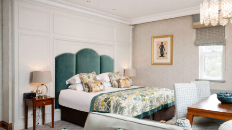 Elegant hotel suite with plush green headboard, floral bedding, large window, chic chandelier, and cosy sitting area.