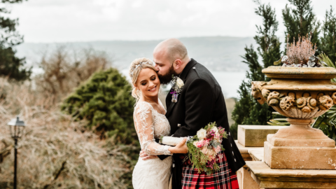 A bride and groom embrace outdoors; the groom, wearing a kilt, kisses the bride's head as she smiles and holds a bouquet. Trees and a stone planter are in the background.