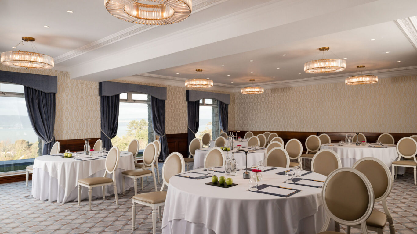 Elegant hotel banquet room with round tables, plush chairs, scenic window views, and luxurious lighting. Ideal for events.