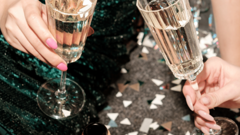 Two people in sparkly attire hold glasses of champagne over confetti-covered floor, with an opened bottle of champagne nearby.