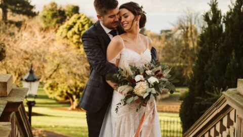 A bride in a white gown holding a bouquet stands on outdoor steps, smiling at the groom who stands behind her; trees and distant water are visible in the background.