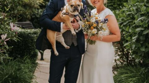 A man in a suit and a woman in a white wedding dress stand outdoors, smiling at the camera. The man holds a small dog, and the woman holds a bouquet of flowers.