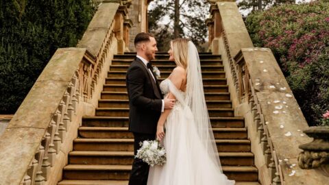 A bride and groom stand facing each other on outdoor stone steps, surrounded by greenery, with the bride holding a bouquet and wearing a long veil.