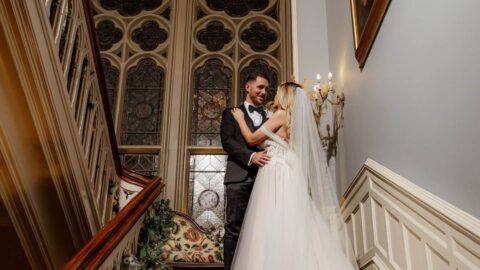 A bride and groom stand together on a grand staircase, embracing in front of a large stained glass window inside an elegant building.