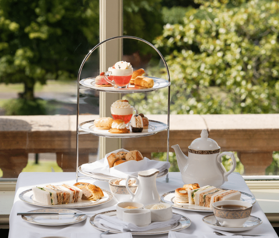A table set for afternoon tea with sandwiches, pastries, and scones on a tiered stand, teapot, cups, and saucers, with a garden view in the background.