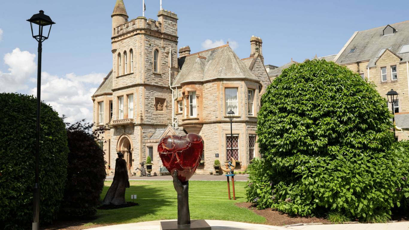 A large red heart sculpture on a pedestal stands in front of a historic stone building with a turret and well-manicured greenery.