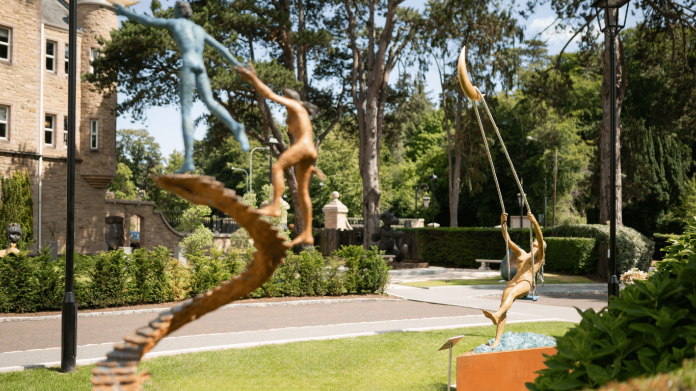 Outdoor view of bronze sculptures depicting human figures reaching for crescent moons, set on a grassy area near a building and surrounded by trees.