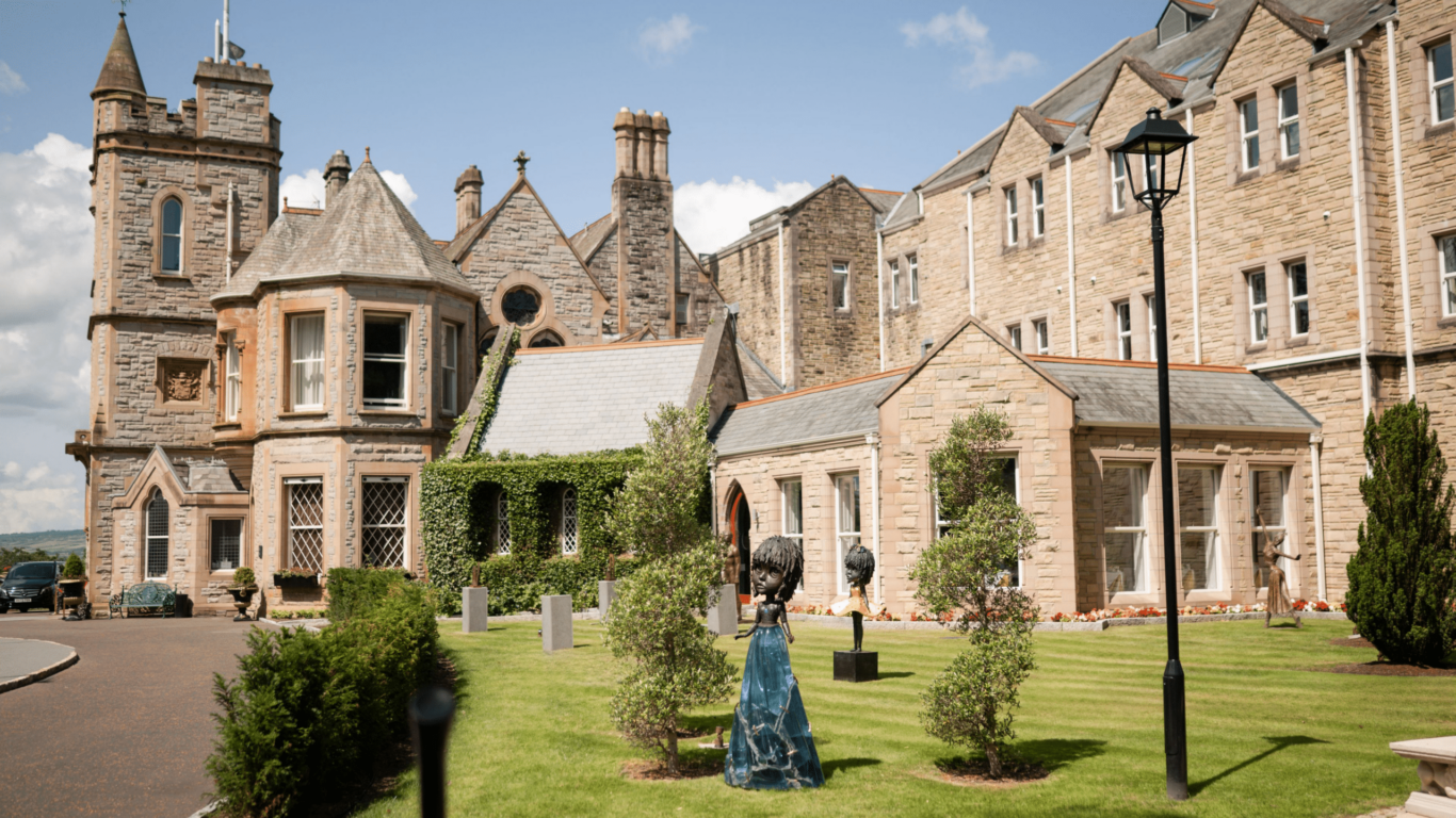 A large historic stone building with multiple gables, turrets, and a well-maintained lawn, featuring sculptures and neatly trimmed bushes in front.