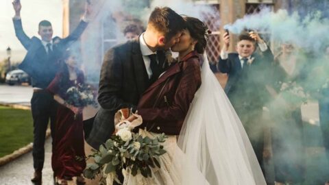 A bride and groom kiss outdoors, holding a bouquet, while guests in formal attire celebrate behind them with colorful smoke bombs. A large building is in the background.