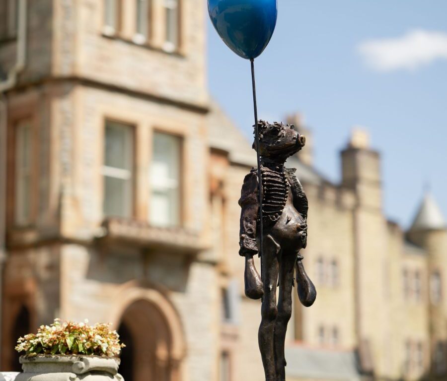 A bronze statue of a child holding a blue balloon stands on a pedestal outdoors in front of a historic stone building.