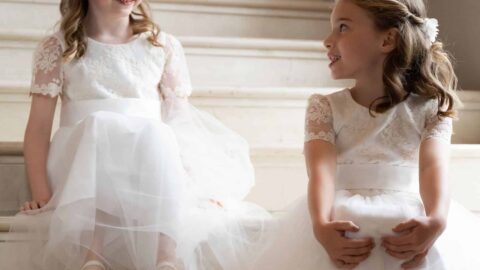 Two young girls in white dresses sit on marble stairs, facing each other and smiling.