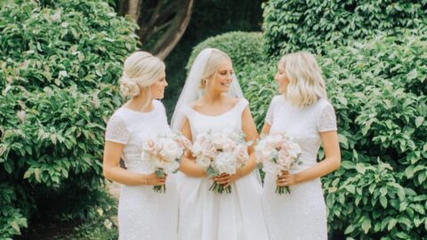 Three women in white dresses, holding bouquets, stand together on a garden path surrounded by greenery. The woman in the center wears a bridal gown and veil.