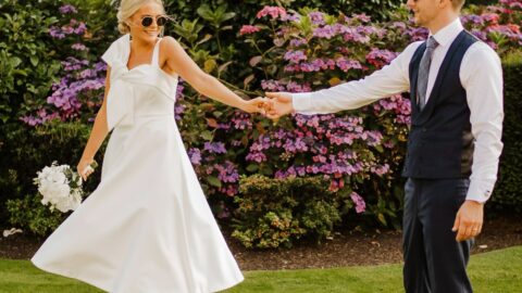 A bride in a white dress and groom in a suit stand on grass, holding hands, with flowering bushes and greenery in the background. Both wear sunglasses.