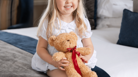 A young girl with blonde hair and a pink bow sits on a bed, smiling and holding a teddy bear with a red ribbon.