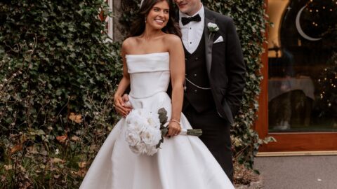 A bride in a strapless white gown holds a bouquet and stands beside a groom in a black tuxedo, posing outdoors by an ivy-covered wall and arched wooden door.