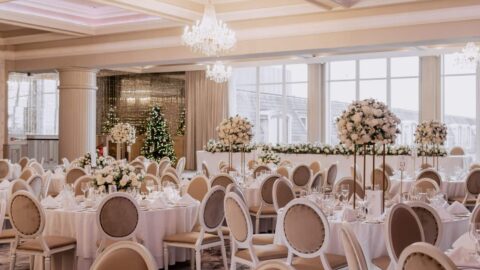 Elegant banquet hall with round tables, beige chairs, white floral centerpieces, chandeliers, and large windows; set up for a formal event or wedding reception.