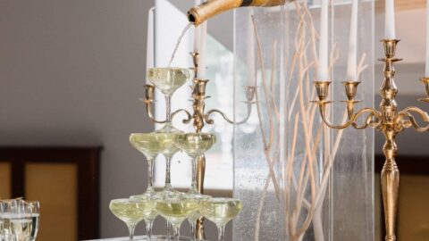 A large champagne bottle pours champagne into a stacked tower of glasses, with additional drinks and gold candelabras on the table below.