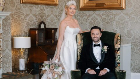 A bride in a white gown and crown stands beside a seated groom in a black tuxedo; they pose in an ornate room with patterned wallpaper and framed portraits.