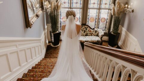 A bride in a long white veil descends a carpeted staircase with stained glass windows and ornate decor in the background.