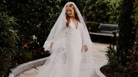 A bride in a long white gown and veil walks along a stone path surrounded by greenery in a garden.