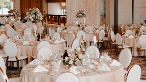 Elegant banquet hall set up for an event with round tables, white chairs, floral centerpieces, glassware, and chandeliers hanging from the ceiling.