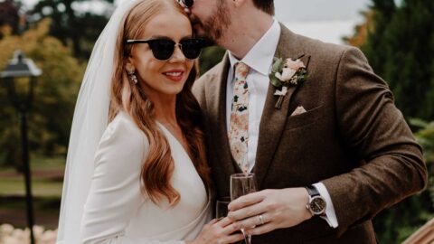 A bride and groom stand outdoors on steps; the groom kisses the bride’s head. Both wear sunglasses and hold glasses, with trees and a cloudy sky in the background.
