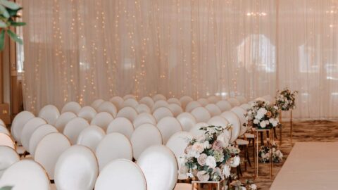 A wedding ceremony setup with rows of white chairs, floral arrangements, chandeliers, and a white aisle in an elegantly decorated indoor venue.