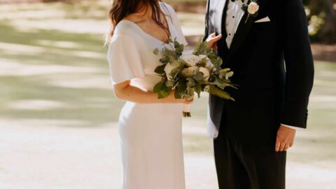 A woman in a white dress holding a bouquet stands next to a man in a black tuxedo outdoors, both facing each other in a sunlit garden setting.