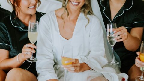 Three women sit on a bed in pajamas, holding glasses of drinks and smiling; the center woman wears a white robe and veil, suggesting she is the bride.