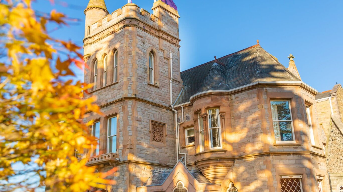 A stone building with a turret and arched windows is shown on a sunny day, with yellow autumn leaves in the foreground and a purple flag on the roof.