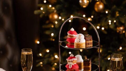 A two-tiered tray with assorted pastries and desserts, flanked by glasses of champagne, a teapot, and a poinsettia, with a decorated Christmas tree in the background.