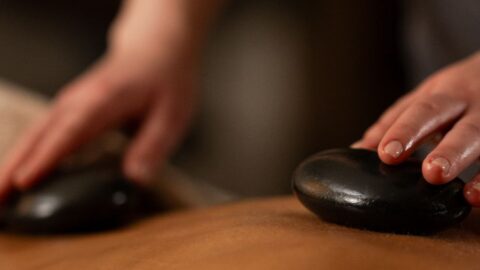 A person receiving a hot stone massage; two smooth black stones are placed on their bare back while a practitioner uses their hands.