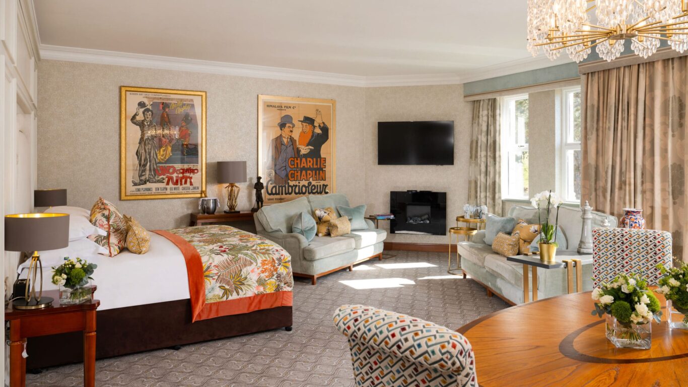 A spacious, well-lit hotel room with a large bed, seating area, wall-mounted TV, vintage posters, chandelier, and floor-to-ceiling windows with curtains.