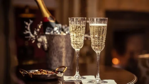 Two filled champagne flutes on a round wooden table with a bowl of snacks and a champagne bottle in an ice bucket; a lit lamp and fireplace are in the background.