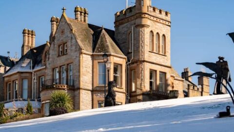 A historic stone building with a turret stands behind a snow-covered hill under a clear blue sky.