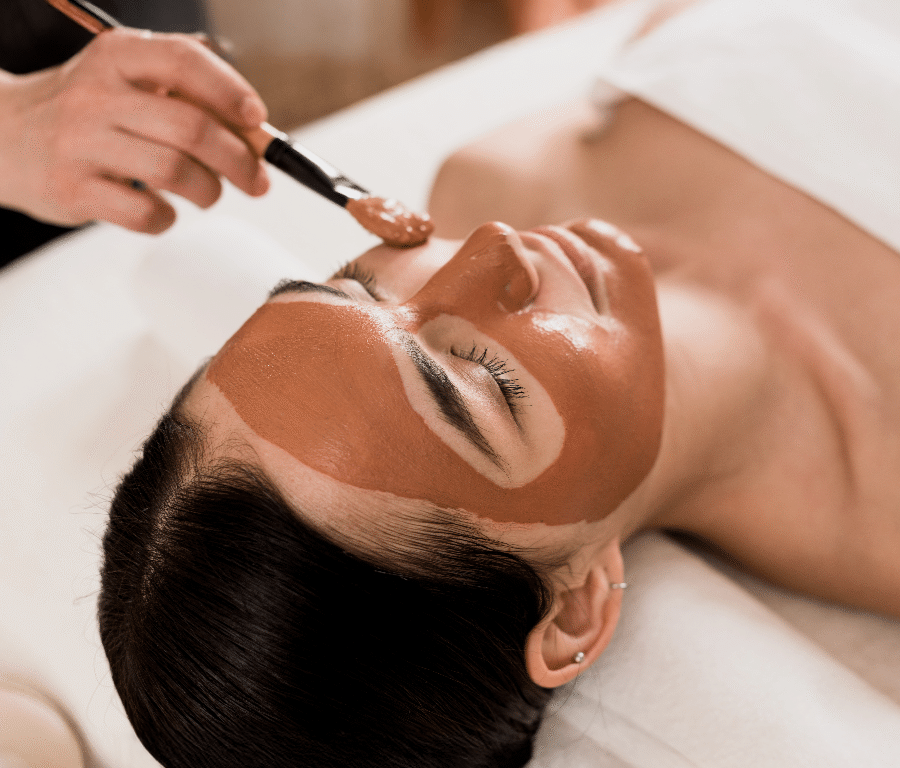 A person lying on a spa bed is having a brown facial mask applied to their face with a brush by another person.