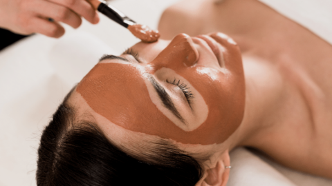 A person lying on a spa bed is having a brown facial mask applied to their face with a brush by another person.