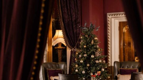 A decorated Christmas tree stands in the center of an elegant lounge area with ornate chairs and a lamp, framed by partially drawn, dark red curtains.