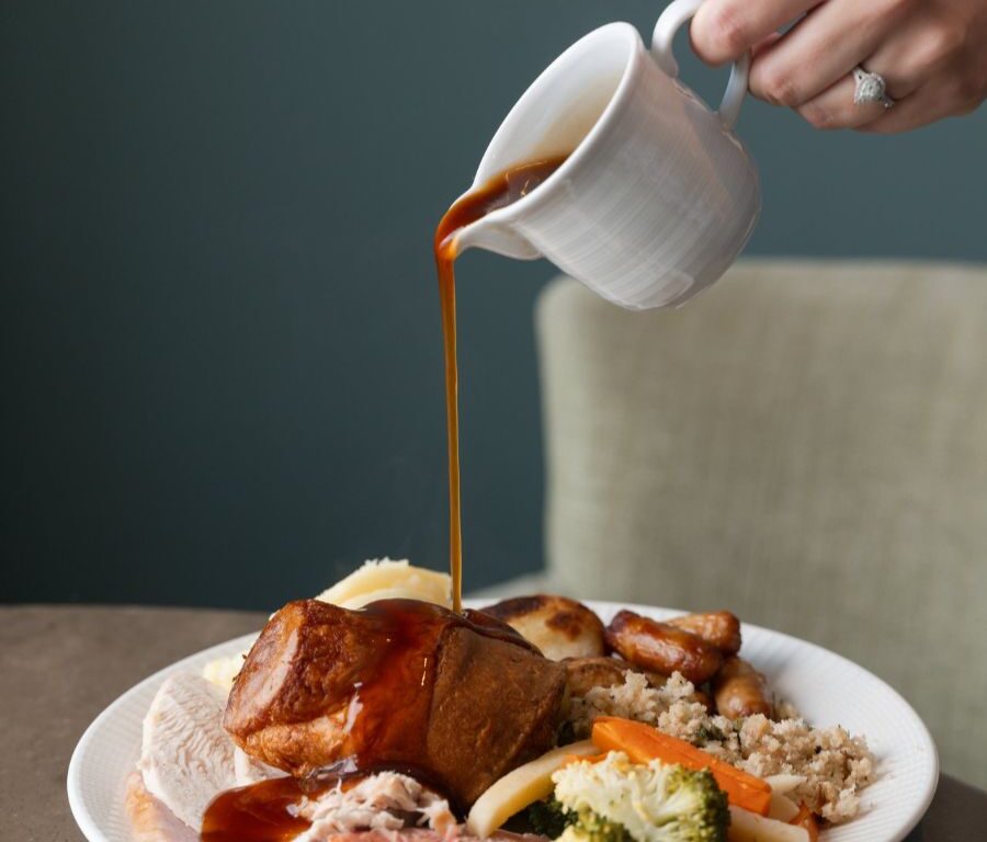 A hand pours gravy from a white jug onto a plate of roast dinner with meats, Yorkshire pudding, potatoes, and vegetables.
