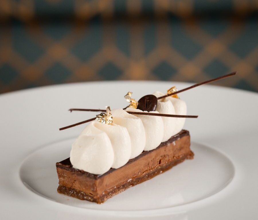 A rectangular chocolate dessert with a layered base, topped with dollops of cream, gold leaf, a cherry, and decorative chocolate sticks, served on a white plate.
