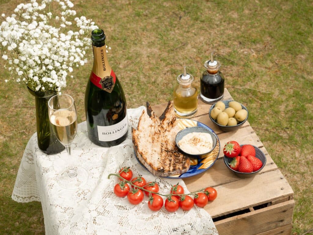 Elegant picnic setup with champagne, fresh strawberries, tomatoes, olives, and grilled toast on a rustic wooden table outdoors.