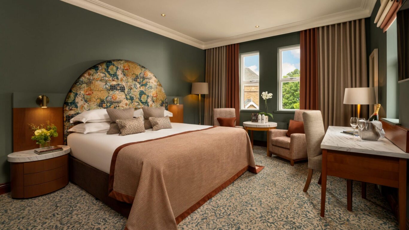 Elegant hotel room with plush king bed, floral headboard, cosy chairs by sunny window, and modern amenities for a relaxing stay.