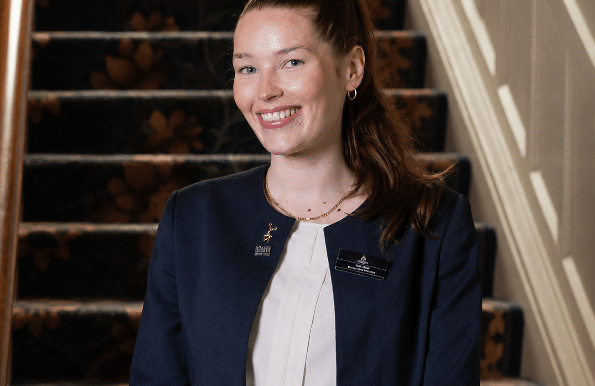 Friendly hotel staff in navy uniform standing by elegant, carpeted staircase, offering a warm welcome to guests.