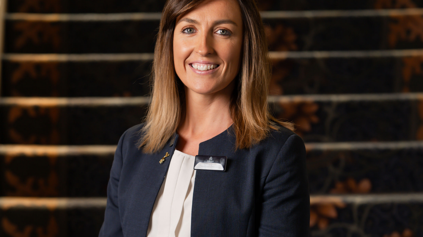 Friendly hotel staff member in smart uniform by elegant carpeted staircase, welcoming guests with a smile.