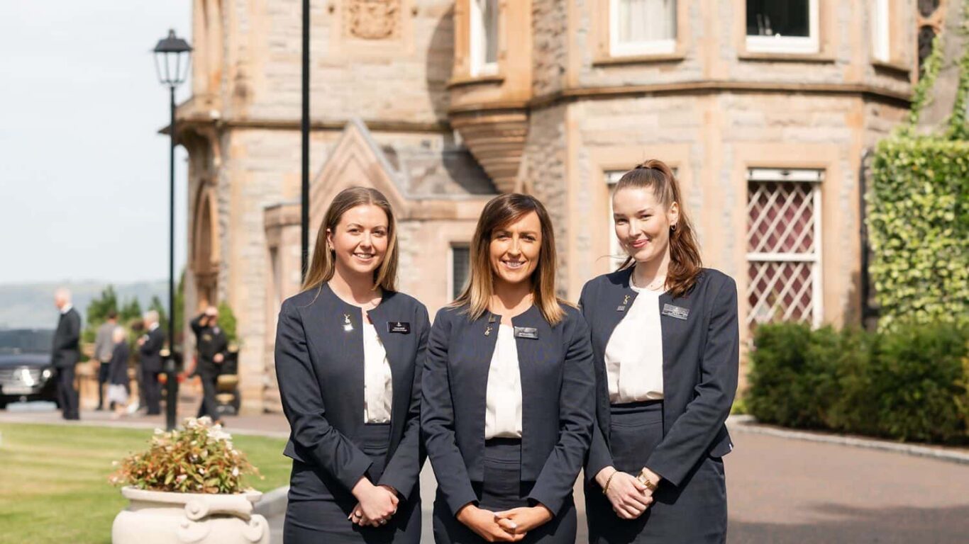Friendly hotel staff stand smiling in front of elegant stone manor with well-kept gardens and scenic views, welcoming guests.