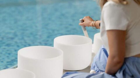 Woman playing crystal singing bowls by serene hotel pool, creating a relaxing atmosphere for guests seeking wellness.