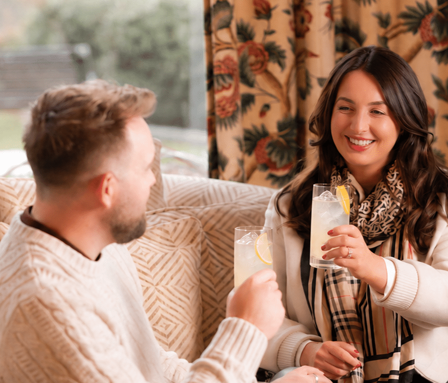 Couple toasting lemon cocktails on a cosy patterned sofa in a hotel lounge, floral curtains and park view visible — relaxing bar and lounge atmosphere